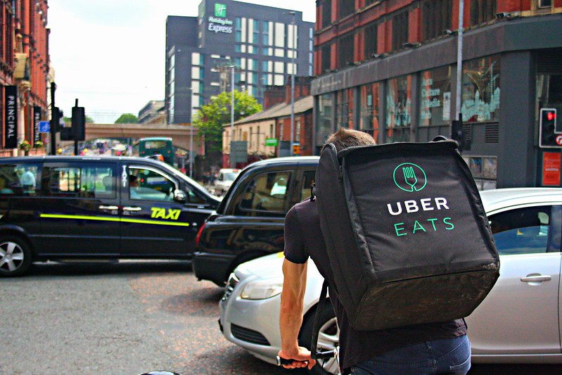 A cyclist wearing an uber eats bag waits at a crossing while taxis drive past